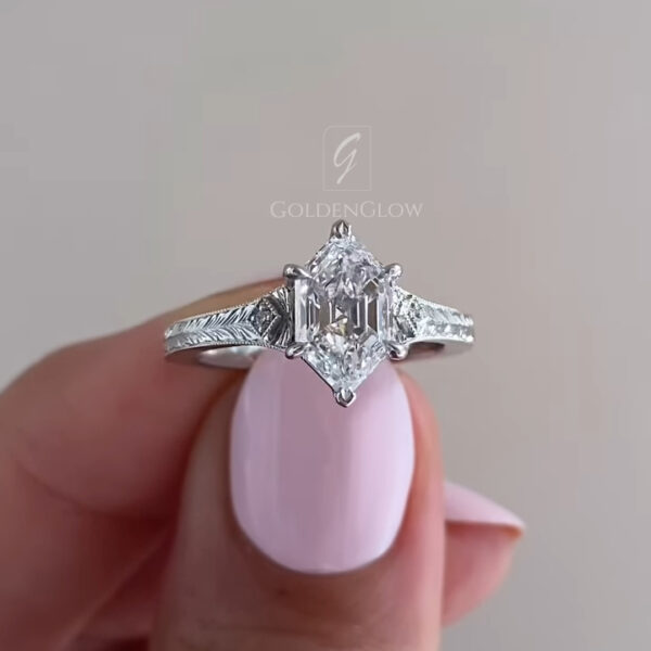 An angled close-up capturing the sophisticated luster of the hexagonal step-cut stone. The lab grown diamond or Moissanite features a large open table that draws the eye deep into the gem. The vintage-inspired white gold setting includes kite accents and milgrain borders that frame the center stone beautifully. The light hits the parallel facets, creating subtle, elegant flashes of white light. This shot showcases the unique silhouette and the meticulous detailing of the antique-style mounting.