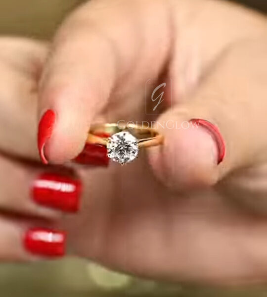 A glamorous angled close-up of a classic solitaire ring worn on a hand with bright red nails. The 18k yellow gold band is polished and slender, leading up to a high-set round brilliant cut lab grown diamond or Moissanite. The gemstone catches the light, displaying a fiery mix of white and rainbow sparkles. This image captures the warmth of the gold against the skin and the commanding presence of the center stone, ideal for a bride seeking a traditional yet bold look.