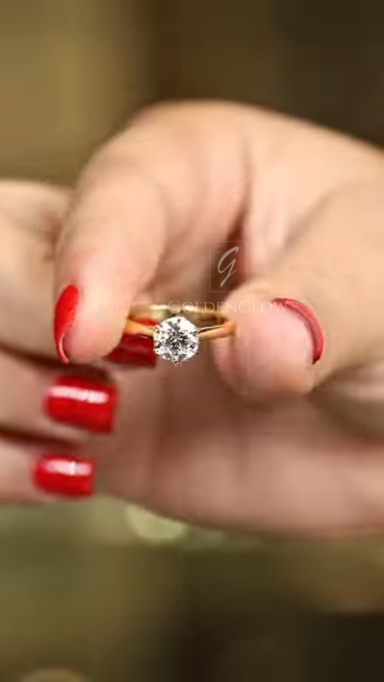 A glamorous angled close-up of a classic solitaire ring worn on a hand with bright red nails. The 18k yellow gold band is polished and slender, leading up to a high-set round brilliant cut lab grown diamond or Moissanite. The gemstone catches the light, displaying a fiery mix of white and rainbow sparkles. This image captures the warmth of the gold against the skin and the commanding presence of the center stone, ideal for a bride seeking a traditional yet bold look.