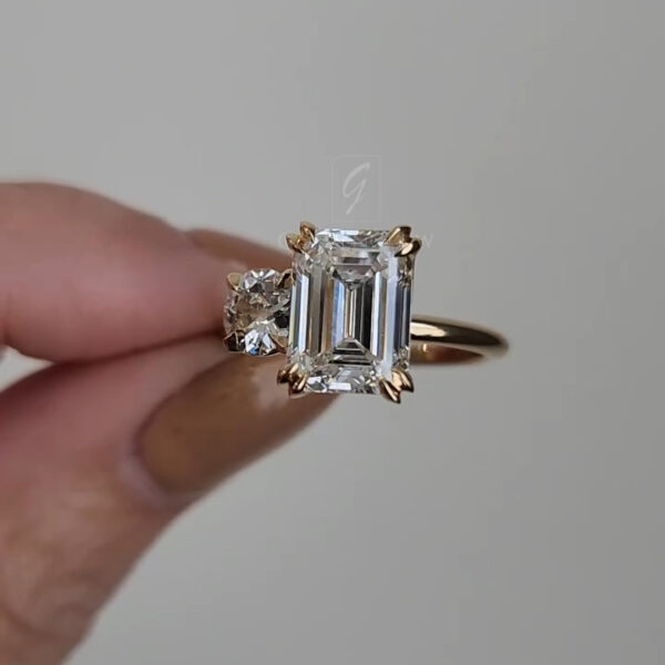 Detailed macro image of the emerald-cut center stone viewed slightly from above. The vertical facet lines within the gemstone are clearly visible, producing a hall-of-mirrors effect. The adjacent round diamond catches light with a bright, scintillating sparkle. The yellow gold prongs and band frame the stones warmly, with smooth metal surfaces reflecting subtle highlights from the soft lighting.