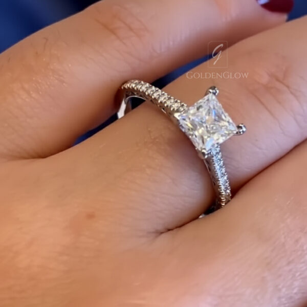 High-resolution image of a square princess-cut ring displayed on a hand, highlighting the refined pavé band and expertly set center stone. The sparkle of the stone catches the light from multiple angles, emphasizing precision craftsmanship and modern elegance. This engagement ring can be made with a lab grown diamond or a moissanite diamond, offering a responsible and affordable alternative to mined diamonds with exceptional brilliance.