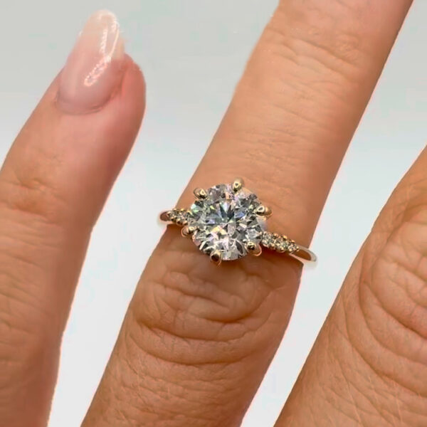 Close-up side view of a yellow gold engagement ring resting on a finger, showcasing a round brilliant center stone held in a six-prong setting. The elevated basket reveals intricate metalwork beneath the stone, while pavé accents shimmer along the band. Designed to highlight sparkle and craftsmanship, this ring is ideal for comparing a lab grown diamond and moissanite diamond, emphasizing brilliance, fire, and refined detail in a silhouette.