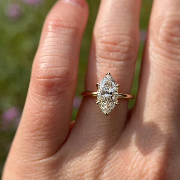 A close-up view of the underside of a gold engagement ring held against a blurred green background. The focus is on the intricate floral-inspired basket setting, which resembles blooming petals cradling the stone. The polished yellow gold band curves elegantly to meet the sophisticated mounting. This exquisite jewelry piece could feature a sustainable lab grown diamond or a sparkling moissanite diamond, symbolizing eternal love. The lighting highlights the warm metal tones and the detailed craftsmanship of the unique petal prongs.
