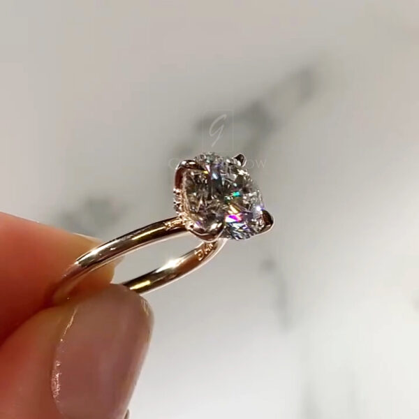 Three-quarter side angle of the diamond ring, showing the transition from the gold band into the sculpted prongs. The round brilliant diamond displays crisp facet reflections with hints of colorful fire. The prongs curve upward in an elegant floral form, securely holding the stone. The background remains neutral and softly diffused, ensuring the ring’s details, shine, and setting architecture remain the focal point.