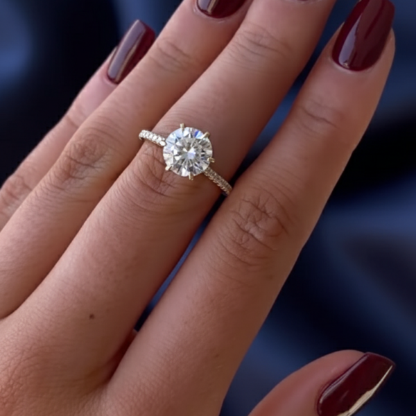 Close-up of a hand wearing a round solitaire engagement ring on a slender finger, set in yellow gold with a delicate pavé band. The lab grown diamond catches the light with a bright star-like sparkle against a soft blue fabric background, highlighting its clarity and brilliance. The image showcases fine craftsmanship, smooth prongs, and elegant proportions while offering a visual comparison often searched alongside moissanite diamond styles for modern engagement rings.