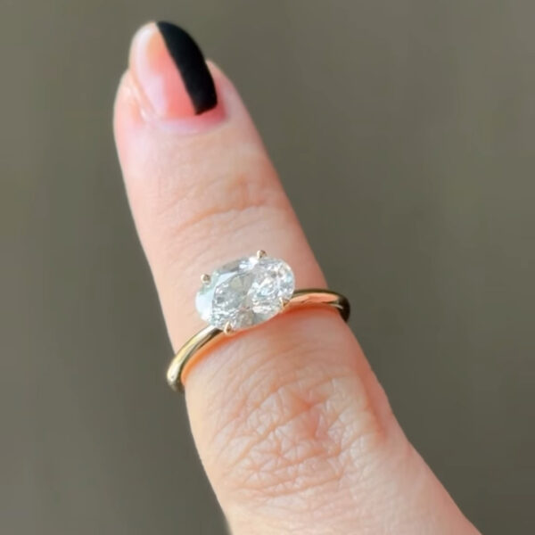 Close-up side profile of an oval diamond solitaire ring, focusing on the prongs and basket setting as it rests on a finger. The yellow-gold band appears slim and refined, complementing the diamond’s brilliance. Light reflections along the stone’s edge highlight craftsmanship and quality. The natural green backdrop keeps attention on the ethical diamond and classic engagement ring silhouette.