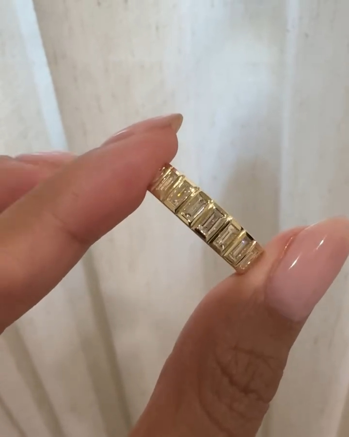 Close-up of a hand wearing a yellow gold eternity-style ring on the ring finger, featuring a continuous row of vertically set baguette-cut diamonds. The diamonds are channel-set, creating a clean, geometric look that reflects light evenly across the band. The ring sits flat against the finger, highlighting its sleek profile and modern design. Natural light emphasizes the clarity of each baguette diamond, while the soft neutral background keeps focus on the refined craftsmanship and polished gold finish.