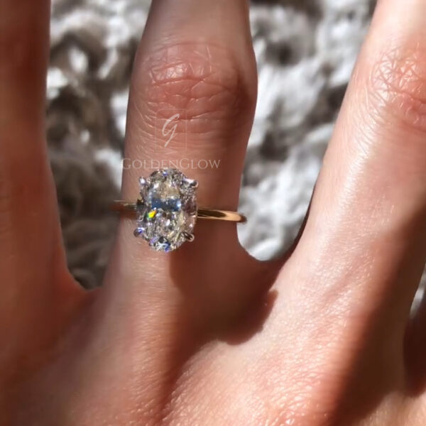 Lifestyle image of an oval solitaire engagement ring worn on the hand against a softly textured background. The oval lab grown diamond displays bright flashes of light, complemented by a polished yellow gold band for a refined, elegant appearance. The clean design focuses attention on the center stone’s brilliance. This engagement ring is also available with a moissanite diamond, providing a sustainable option with exceptional sparkle and durability.