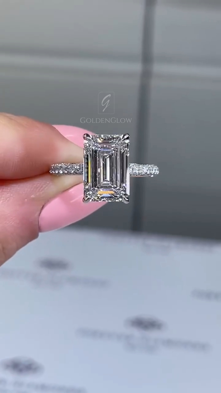 Close-up side profile of a luxury engagement ring featuring an emerald-cut center stone set in a white metal band with pavé diamonds. The elevated basket and prong structure highlight the stone’s depth and clean step facets. This detailed view showcases craftsmanship and sparkle, ideal for comparing a lab grown diamond versus a moissanite diamond in terms of brilliance, fire, and setting height on a modern fine jewelry ring.