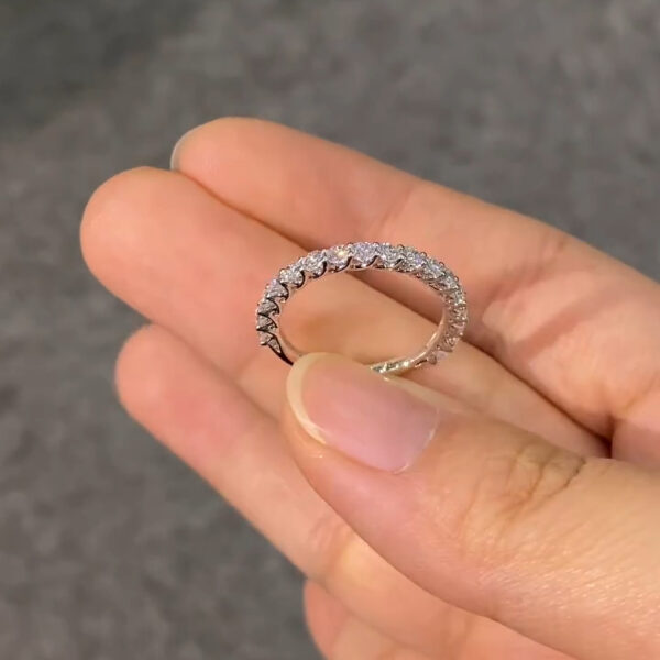 Close-up view of a slim eternity ring held between fingers against a soft gray fabric background. The circular band is set with closely spaced, round-cut stones that reflect light evenly around the entire ring. The polished metal appears white-toned and smooth, with prong settings visible along the outer edge. The image highlights sparkle and craftsmanship, commonly associated with a lab grown diamond or moissanite diamond eternity band designed for everyday wear or stacking.