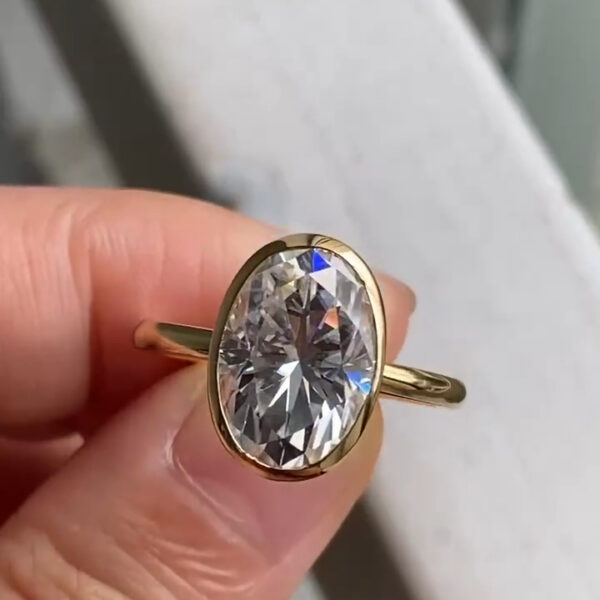 Close-up of an oval bezel-set engagement ring held between fingers, showcasing a brilliant lab grown diamond with crisp facets and bright white sparkle. The polished yellow gold band frames the stone smoothly, reflecting light along the bezel edge. This elegant design highlights the clarity and fire often compared between a lab grown diamond and a moissanite diamond, making it ideal for modern, minimalist engagement ring styles.