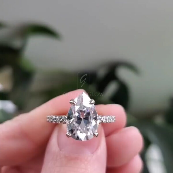 Close-up image of a pear-shaped clear gemstone engagement ring held gently between fingers. The ring features a polished precious metal band with pavé-set accent stones along the shoulders. The side profile highlights the elevated prong setting that secures the pear-cut center stone, allowing light to pass through and enhance sparkle. The background is softly blurred with neutral tones, keeping focus on the craftsmanship, stone shape, and band detailing.