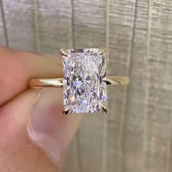 Close-up of a rectangular radiant-cut lab-grown diamond or moissanite engagement ring held between fingers, featuring a bright elongated stone secured by four gold prongs. The yellow gold band has a smooth polished finish that reflects soft ambient light. The gemstone displays crisp facet definition, strong white brilliance, and subtle rainbow flashes, with a neutral textured background emphasizing the clarity, ethical origin, and modern elegance of the lab-created center stone.