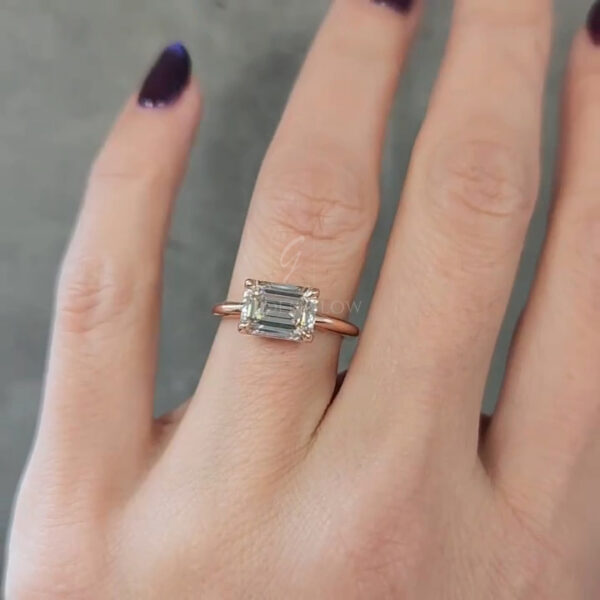 Close-up view of a hand wearing an emerald-cut diamond ring on the finger. The rectangular step-cut stone is set horizontally in a minimalist rose gold band with four prongs securing each corner. The diamond’s clean facets reflect soft light, emphasizing clarity and symmetry. The slim band sits flush against the finger, creating an elegant, understated look. The background is softly blurred gray, keeping attention focused on the ring’s geometry and refined craftsmanship.