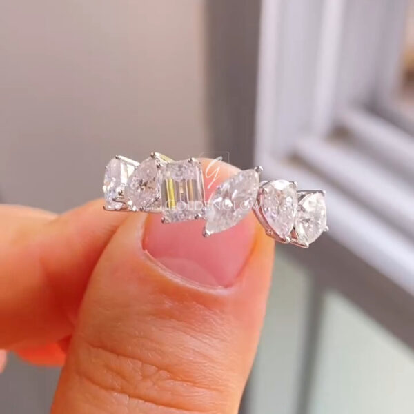 Close-up view of a multi-stone diamond band held between fingers, featuring a symmetrical arrangement of mixed-cut diamonds. An emerald-cut diamond sits at the center, flanked by marquise and oval-shaped stones, all set in polished white metal. The diamonds catch natural window light, highlighting their clarity and sparkle, while the prong settings and smooth band edges emphasize fine craftsmanship and a modern yet elegant design.