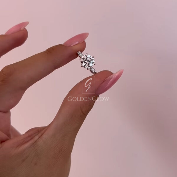 Close-up side profile of a round solitaire engagement ring held between manicured fingers with glossy pink nails. The ring features a brilliant lab grown diamond secured in a classic prong setting, paired with a slim pavé band that enhances sparkle from every angle. Crafted to resemble fine luxury jewelry, this design is also available with a moissanite diamond option, offering an ethical and radiant alternative with exceptional fire and brilliance.