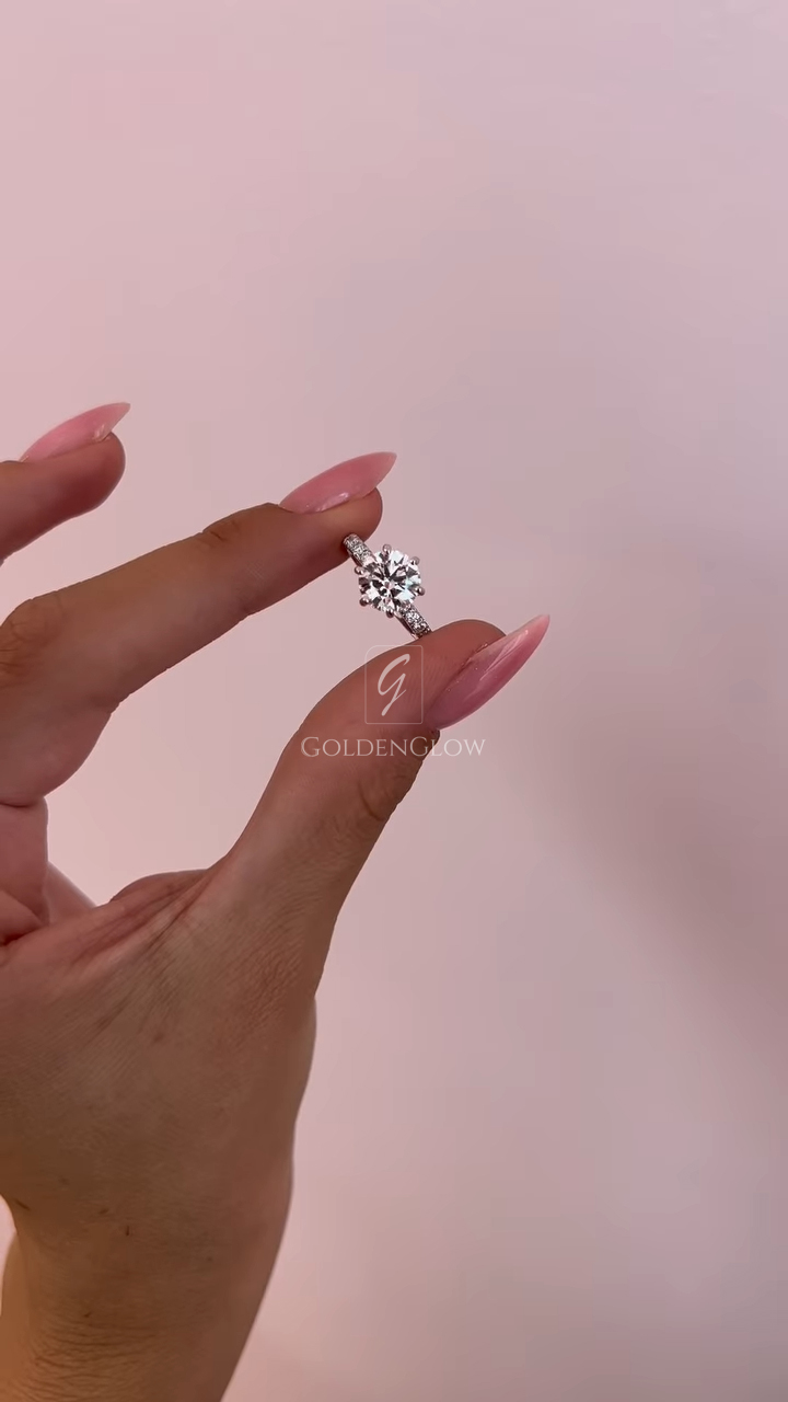 Close-up side profile of a round solitaire engagement ring held between manicured fingers with glossy pink nails. The ring features a brilliant lab grown diamond secured in a classic prong setting, paired with a slim pavé band that enhances sparkle from every angle. Crafted to resemble fine luxury jewelry, this design is also available with a moissanite diamond option, offering an ethical and radiant alternative with exceptional fire and brilliance.