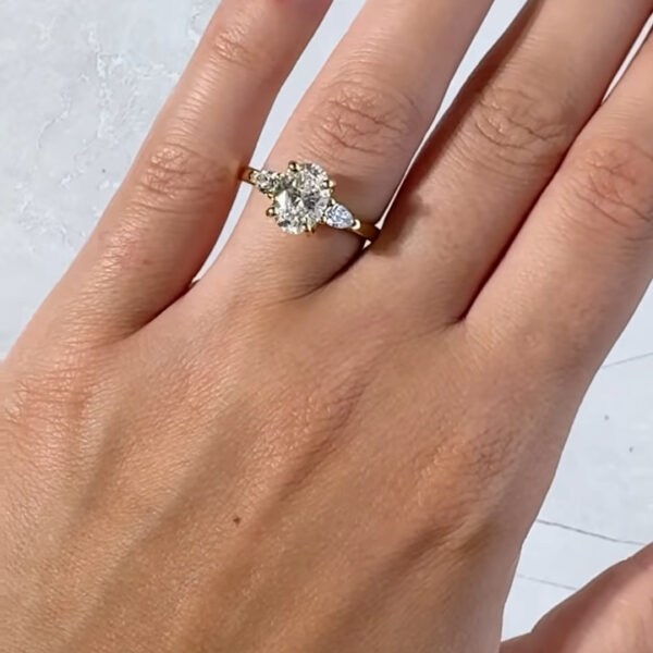 Top view of a three-stone oval engagement ring worn on the hand with soft nude manicured nails. The centerpiece features a brilliant oval lab grown diamond, accented by pear-shaped side stones set in warm yellow gold. The polished band creates a timeless and elegant silhouette. This engagement ring is also available with a moissanite diamond, offering a sustainable, ethical alternative with exceptional sparkle and fire.