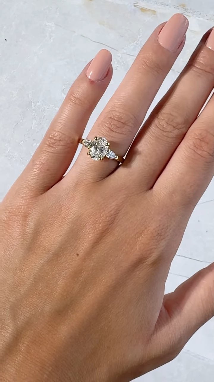 Top view of a three-stone oval engagement ring worn on the hand with soft nude manicured nails. The centerpiece features a brilliant oval lab grown diamond, accented by pear-shaped side stones set in warm yellow gold. The polished band creates a timeless and elegant silhouette. This engagement ring is also available with a moissanite diamond, offering a sustainable, ethical alternative with exceptional sparkle and fire.