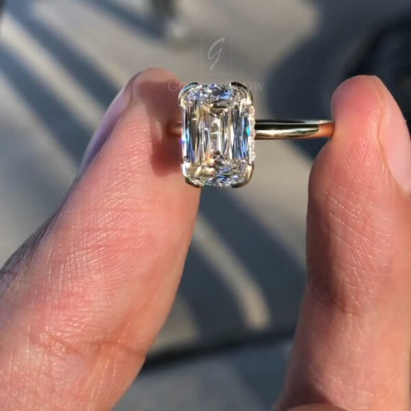 A stunning front view of a massive elongated radiant-cut solitaire ring. The centerpiece is a dazzling lab grown diamond or Moissanite, oriented vertically to elongate the finger. It is secured by a modern yellow gold "clutch" setting that wraps the top and bottom edges. The stone's crushed-ice faceting reflects light beautifully, creating a disco-ball effect. This sustainable luxury piece blends architectural design with the brilliance of a colorless diamond alternative.