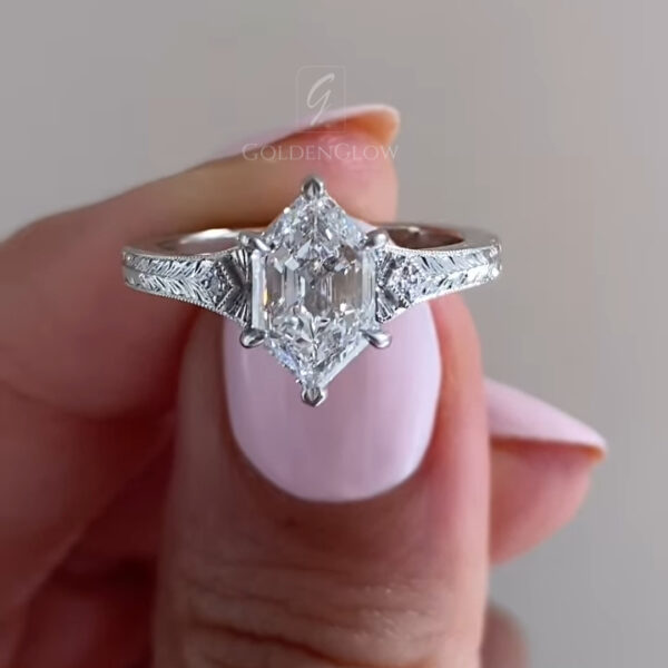 A detailed side profile of a vintage-inspired engagement ring in gleaming white gold or platinum. The band features exquisite hand-engraved wheat patterns with delicate milgrain edges, evoking an Art Deco aesthetic. The center stone, a unique elongated hexagonal step-cut lab grown diamond or Moissanite, is set high in a basket to maximize light capture. The prongs are sharp claws. This view highlights the artistry of the antique-style finish and the contrast between the textured exterior and polished interior.