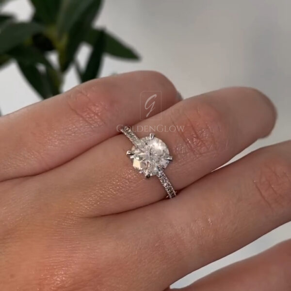 A stunning side-profile view of a handcrafted oval halo engagement ring held between fingertips. This angle reveals the sophisticated architecture of the setting, where the main oval-cut lab grown diamond or Moissanite rests high in a basket. The image highlights the "cathedral" style shank that rises to meet the halo, adding structural elegance. The band is lined with micro-pavé stones that glitter even from the side. You can clearly see the depth of the center stone and the delicate metalwork supporting the halo.