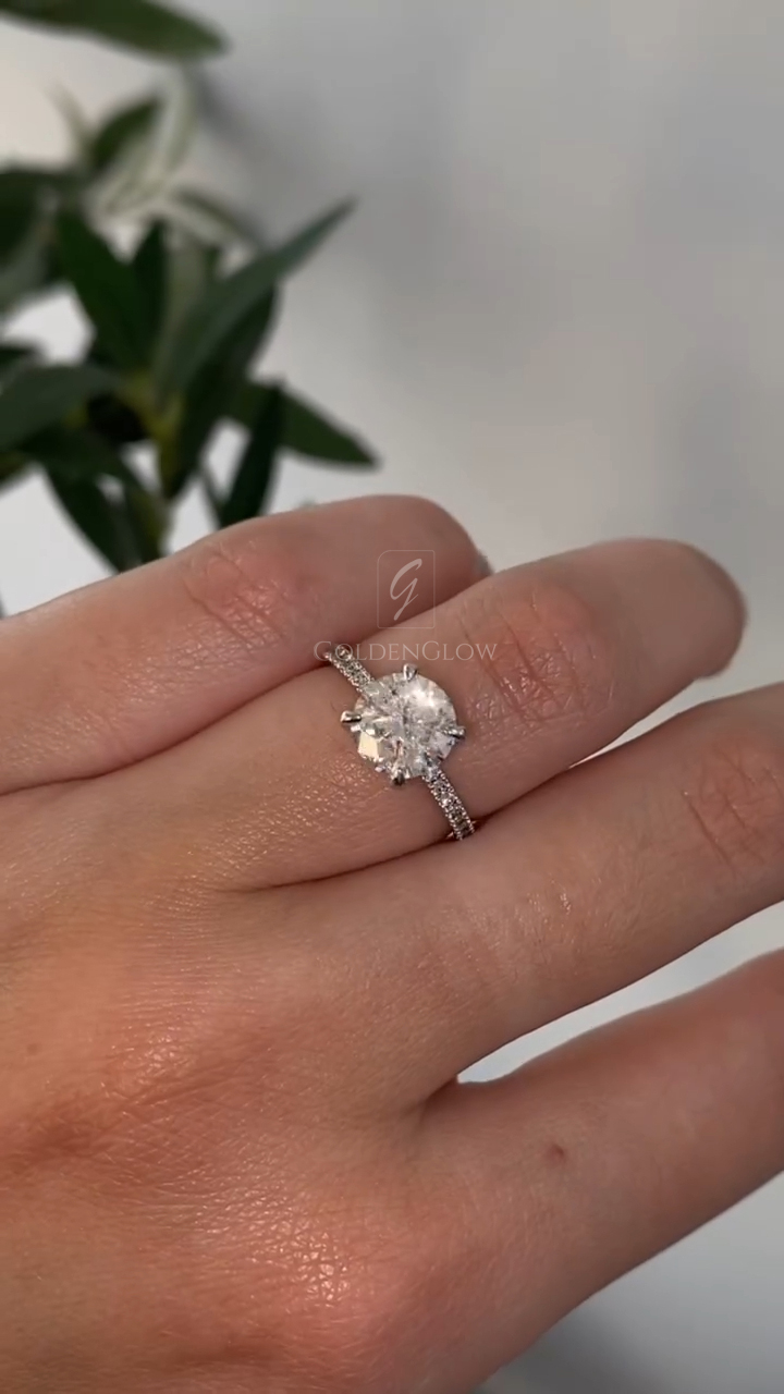 A stunning side-profile view of a handcrafted oval halo engagement ring held between fingertips. This angle reveals the sophisticated architecture of the setting, where the main oval-cut lab grown diamond or Moissanite rests high in a basket. The image highlights the "cathedral" style shank that rises to meet the halo, adding structural elegance. The band is lined with micro-pavé stones that glitter even from the side. You can clearly see the depth of the center stone and the delicate metalwork supporting the halo.