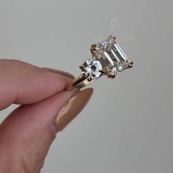 Close-up view of a yellow gold engagement ring held between fingers against a light gray background. The ring features a large emerald-cut clear gemstone set vertically in a four-prong setting, with a smaller round brilliant diamond accent positioned to one side. The gemstones reflect light sharply, showing crisp step facets on the emerald cut and bright sparkle on the round stone. The polished gold band appears smooth and slightly tapered near the setting.