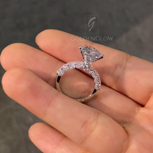 Rear view of a round engagement ring resting on an open hand, featuring a brilliant diamond secured in a raised prong setting. The band is adorned with round accent stones that curve along the top half of the ring, adding layered sparkle and dimension. This image highlights the clean brilliance of a lab grown diamond while allowing visual comparison to the stronger fire often seen in a moissanite diamond under natural outdoor lighting.