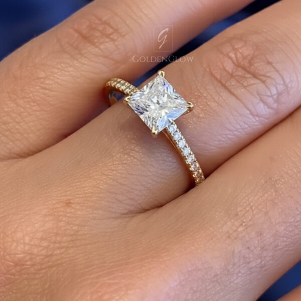 Close-up image of a square princess-cut engagement ring worn on a hand, featuring a brilliant center stone set in a delicate pavé band. The ring sparkles under soft lighting against a blue fabric background, highlighting the crisp facets and elegant prongs. This ring design is available with a lab grown diamond or a moissanite diamond, offering a sustainable and ethical alternative to traditional mined stones while maintaining exceptional brilliance and clarity.
