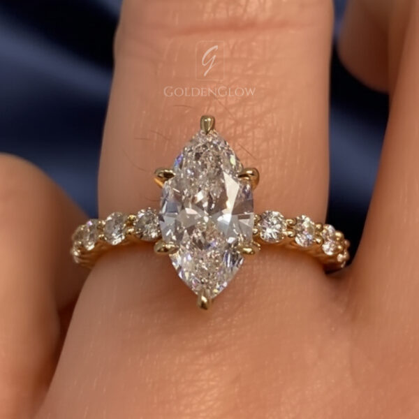 Close-up of a marquise-cut engagement ring worn on a hand against a deep blue satin background. The center stone is elongated and set vertically in yellow gold prongs, with a delicate pavé band adding extra sparkle along the shank. Fine facets reflect light softly across the surface, highlighting clarity and brilliance. This image showcases the visual appeal often compared between a lab grown diamond and a moissanite diamond, emphasizing elegance, fire, and refined craftsmanship.