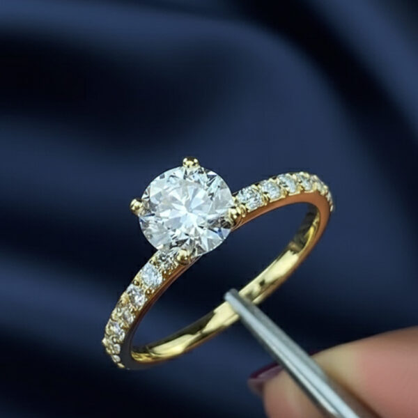 Close-up of a yellow gold engagement ring held with precision tweezers, featuring a round brilliant solitaire set in a four-prong setting with a pavé diamond band. The center stone sparkles against a soft navy satin background, highlighting fine craftsmanship and elegant proportions. This image is ideal for showcasing a lab grown diamond alternative compared visually with a moissanite diamond, emphasizing clarity, brilliance, and luxury jewelry design details.