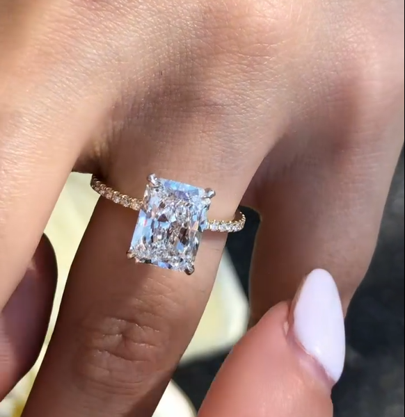 Close-up view of a rectangular radiant-cut engagement ring worn on a hand with deep burgundy nails, set on a slim pavé band. The center stone flashes crisp white brilliance and rainbow fire under natural light against a soft blue fabric background. This image highlights the elegant proportions and sparkle often compared between a lab grown diamond and a moissanite diamond, showing clarity, facet definition, and refined craftsmanship in fine jewelry design.