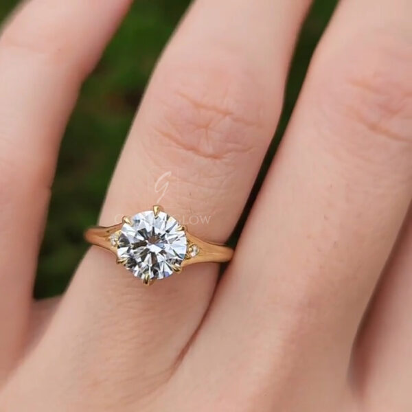 Side-angle close-up of a yellow gold solitaire engagement ring on a finger, showing the diamond slightly tilted. The prongs and open gallery are visible beneath the stone, highlighting craftsmanship and light performance. The gold band curves gracefully, maintaining a slim profile. Natural outdoor lighting and a softly blurred green backdrop enhance the diamond’s brilliance while keeping the focus on the ring’s design.
