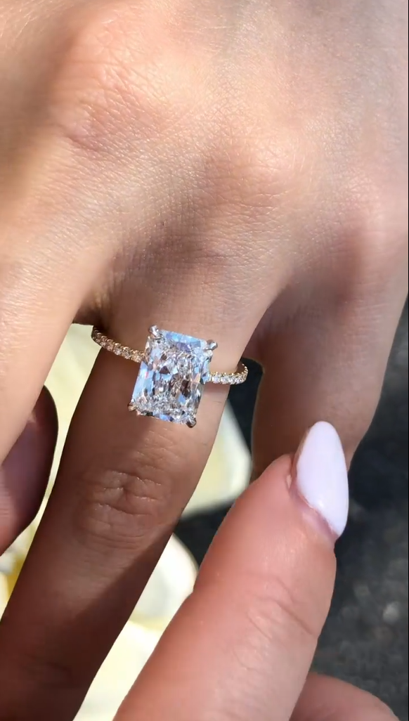 Close-up view of a rectangular radiant-cut engagement ring worn on a hand with deep burgundy nails, set on a slim pavé band. The center stone flashes crisp white brilliance and rainbow fire under natural light against a soft blue fabric background. This image highlights the elegant proportions and sparkle often compared between a lab grown diamond and a moissanite diamond, showing clarity, facet definition, and refined craftsmanship in fine jewelry design.