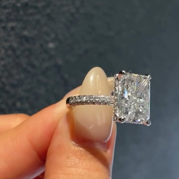 Angled top-down view of a diamond or moissanite ring highlighting the radiant-cut center stone and pavé-set shoulders. The elongated gemstone shows symmetrical facets and exceptional brilliance. The white metal band tapers gently, drawing attention to the center stone. Natural lighting enhances fire and sparkle, making this image ideal for showcasing ethical engagement ring elegance.