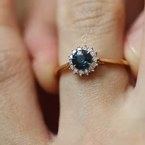Top-down view of a lab-created blue stone ring resting on a finger, showcasing the symmetrical halo of moissanite stones and polished gold band. The image highlights sustainable materials, refined proportions, and a timeless design suitable for engagement or special occasions.