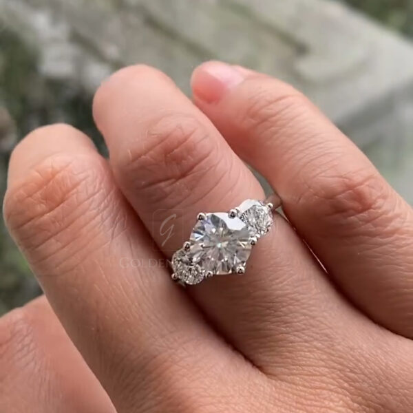 Close-up of a hand wearing a three-stone diamond engagement ring, featuring a round brilliant center diamond flanked by pear-shaped side stones. The ring is set in white metal with delicate prongs, resting naturally on the finger. Soft outdoor light highlights the sparkle of the diamonds, with a blurred stone and greenery background creating an elegant, romantic atmosphere.