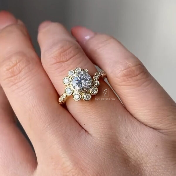 Close-up view of a yellow gold halo engagement ring worn on a hand, featuring a round brilliant-cut center diamond surrounded by a floral-style halo of bezel-set round diamonds. The polished yellow gold band includes small accent diamonds along the shoulders, adding sparkle and texture. The ring is photographed in soft natural light against a neutral gray background, highlighting the contrast between the warm gold metal and the icy brilliance of the diamonds while showing realistic scale and everyday wear.