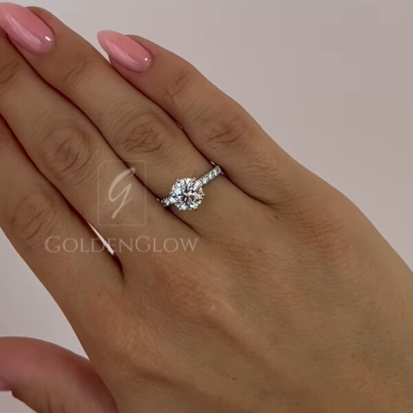 Lifestyle shot of a hand wearing a classic round solitaire engagement ring. The lab grown diamond sits in a timeless prong setting, paired with a slim pavé band that enhances sparkle while remaining elegant and understated. Perfect for everyday wear or special occasions, this ring is also available with a moissanite diamond, offering a sustainable, radiant option with stunning light performance.