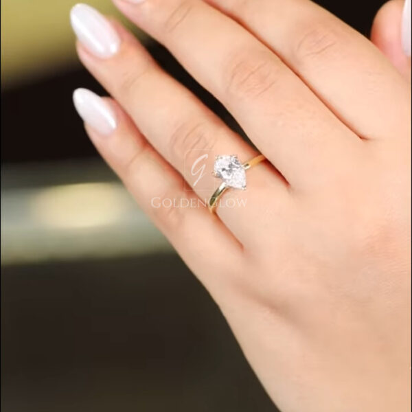 A stunning front view of a pear-shaped solitaire ring held in fingers. The center stone, a premium diamond or Moissanite, features a classic teardrop silhouette with a rounded bottom and a pointed top. It is set in 18k yellow gold with a protective V-tip prong. The stone appears colorless and transparent, shining with internal fire. This image highlights the simplicity of the solitaire design, which lets the unique shape of the gem take center stage.