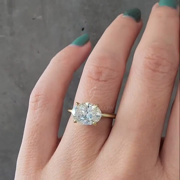 Close-up of a hand wearing an oval-cut clear gemstone solitaire ring set in yellow gold. The oval stone is secured with four yellow gold prongs and sits on a slim, polished band. The ring is photographed from the top, showing the elongated oval shape and faceting of the stone. The hand is positioned against a neutral gray background, allowing the gemstone’s sparkle and proportions to remain the main focus.
