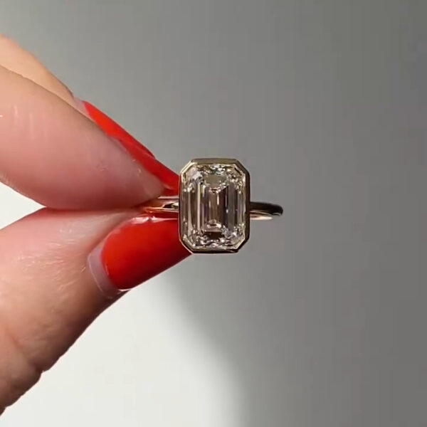 Front-facing close-up of an emerald-cut engagement ring held between fingers with red nail polish, featuring a step-cut diamond set in a sleek bezel-style setting on a yellow gold band. The elongated facets reflect clean, linear flashes under natural light. This image allows visual comparison between the refined brilliance of a lab grown diamond and the higher fire often associated with a moissanite diamond in similar lighting conditions.