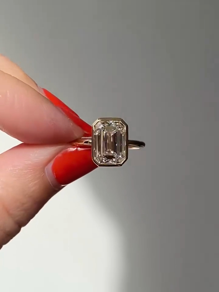 Front-facing close-up of an emerald-cut engagement ring held between fingers with red nail polish, featuring a step-cut diamond set in a sleek bezel-style setting on a yellow gold band. The elongated facets reflect clean, linear flashes under natural light. This image allows visual comparison between the refined brilliance of a lab grown diamond and the higher fire often associated with a moissanite diamond in similar lighting conditions.
