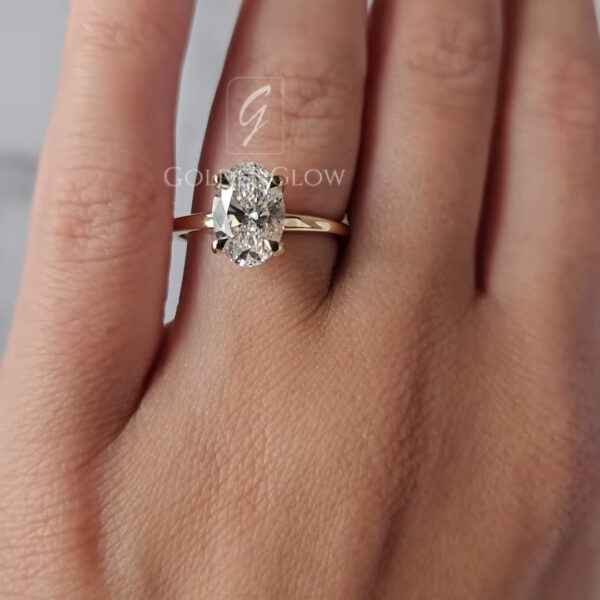 Front-facing view of an oval engagement ring displayed delicately between fingertips against a soft neutral background. The centerpiece oval lab grown diamond reflects bright flashes of light, set in a smooth yellow gold band for a minimalist, refined appearance. Designed for modern elegance and lasting wear, this ring is also offered with a moissanite diamond option, providing a radiant, eco-friendly choice with diamond-like clarity.